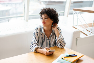 Beautiful cheerful curly woman using mobile phone while sitting in cafe