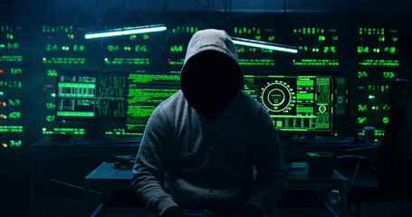 Portrait of  incognito hacker using computer for organizing massive data breach attack on corporate servers. Secret location surrounded by displays, servers and cables.