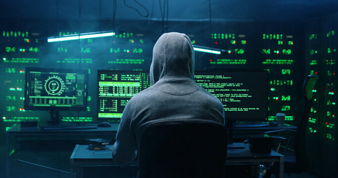 Rear view of incognito hacker using computer for organizing massive data breach attack on corporate servers. Secret location surrounded by displays, servers and cables.