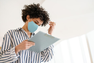 Beautiful woman in face mask holding clipboard and writing down notes