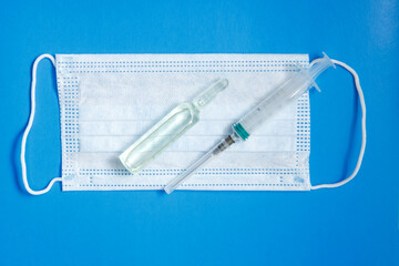 An ampoule with a vaccine, a medical mask and a syringe lie on a blue background. The concept of vaccination against coronavirus. Copy space.