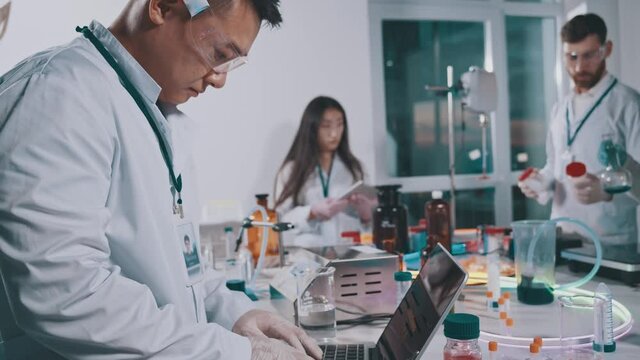 Asian scientist using laptop computer while collaborating in laboratory. Multi-ethnic professional lab workers developing medications in research facility center.