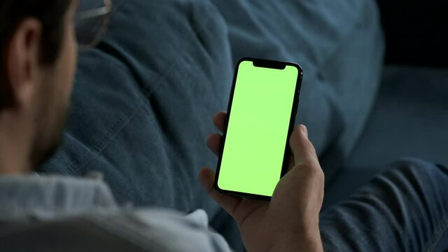 Man lying on couch using smartphone with chroma key green screen at night, scrdoing various gestures like swiping and scrolling - internet, communications concept close up 4k template