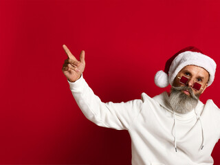 Fototapeta na wymiar Hipster grandpa in a Santa hat, in fashionable sunglasses and with a beard shows a thumbs up, to an empty spot, on a red background.