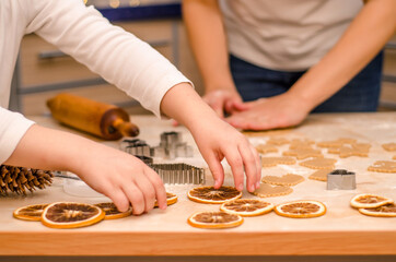 Hands of mother and daughter prepare festive gingerbread cookies for christmas, girl decorates table with dry oranges
