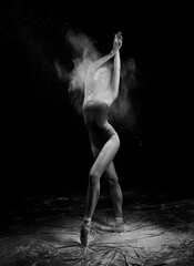 A beautiful slender ballet dancer girl wearing a bodysuit and pointe shoes, posing dancing among the clouds of flying flour on a black background. Artistic, commercial, monochrome design