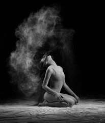 A beautiful slender ballet dancer girl wearing a bodysuit, sensually waves her long hair while sitting on the floor among the clouds of flying flour, on black. Artistic, commercial, monochrome design