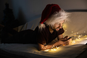Handsome young man lying on the couch wearing santa hat, using his phone and smiling in dark...