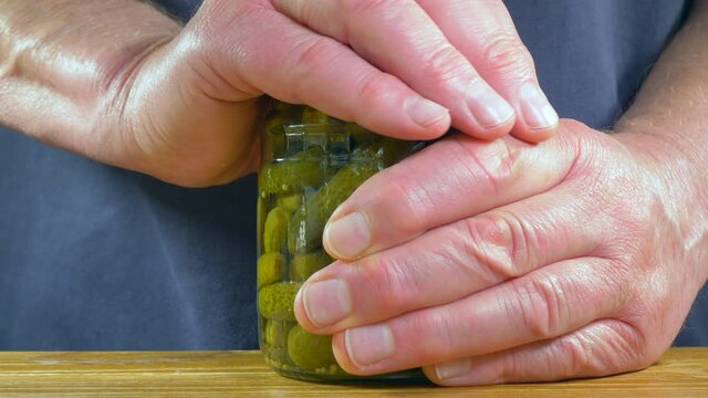Closeup POV shot of a man&rsquo;s hands twisting the lid and removing it from a jar of pickled mini gherkins / cornichons, with red chillis and mustard seeds in vinegar.