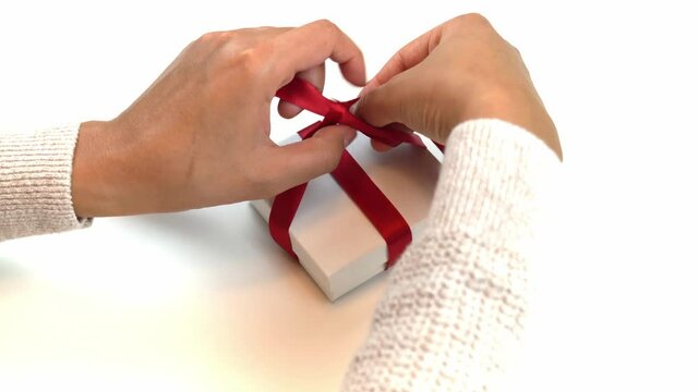 Holiday or Christmas close up of a mixed Race African American woman's hands wrapping and tying a red ribbon bow around a white jewelry gift box present on a white background.