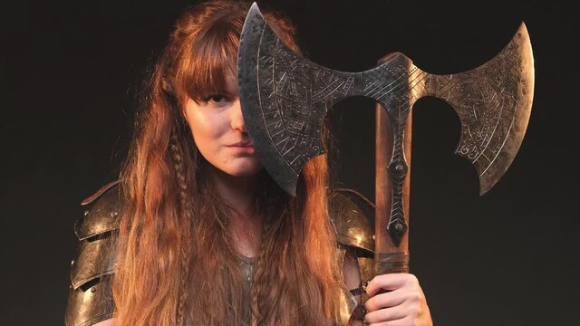 Strong and furious female valkyrie from the north with long brown hairs in antique clothing and armour poses in dark background looking at camera and holding a huge double axe hiding half of her face.