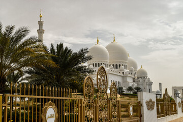 Sheikh Zayed Grand Mosque and its beautiful gate and some palm trees in a cloudy day. Abu Dhabi, United Arab Emirates.