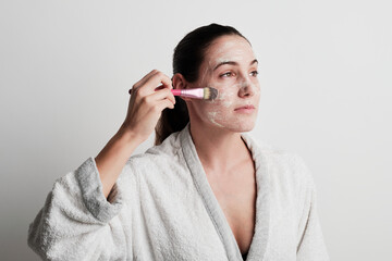 Young woman applying cream in her face with a brush wearing a white bathrobe over a whit background