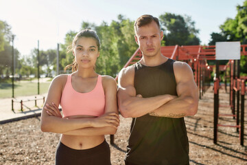 Portrait of muscular young guy and sportive girl looking at camera, standing with arms crossed while exercising together at outdoor gym