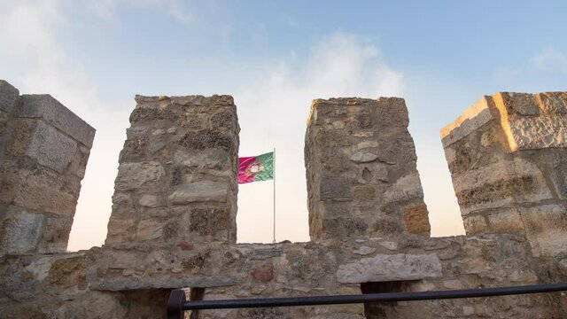 Time lapse of Saint Jeorge castle  during a sunny cloudy day at the sunset. There is the stone bricks medieval wall in close up at the portugal flag in background