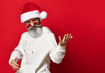 Fototapeta na wymiar stylish and handsome senior hipster wearing a Santa hat winks and beckons for New Year's discounts. Man and Santa hat stands on a red background and looks to the side. Place for text.
