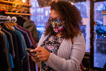 African American mixed woman with curly hair and wearing a protective mask is texting on her smart phone while shopping during Covid-19 pandemic. 