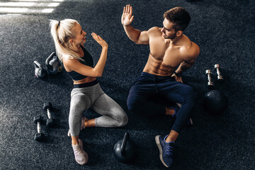 young man and woman sitting on the floor and giving each other five in the gym, fitness people after workout in the gym