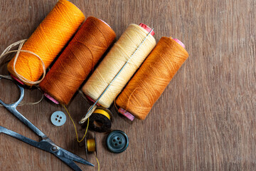 Four different shades of yellow and brown colored sewing thread spools with needle and sewing accessories on brown wooden background.