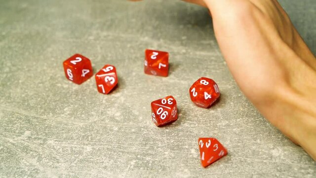 Hand throwing lots of red polyhedral RPG game dice on the table, super slow motion, closeup. Multiple dice throw up close, role playing table top board games, larp, geek, nerd culture abstract concept
