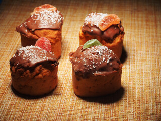 Four homemade cupcakes lie on the tablecloth. Cupcakes are decorated with chocolate icing, coconut chips, and pieces of marmalade.