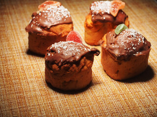 Four homemade cupcakes lie on the tablecloth. Cupcakes are decorated with chocolate icing, coconut chips, and pieces of marmalade.