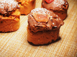 Four homemade cupcakes lie on the tablecloth. Cupcakes are decorated with chocolate icing, coconut chips, and pieces of marmalade.