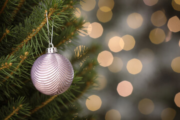 Shiny lilac ball hanging on Christmas tree against festive lights. Space for text