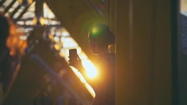 Silhouette young African-American woman with bun takes selfie while standing on modern bridge gallery illuminated bright sunset at backlit