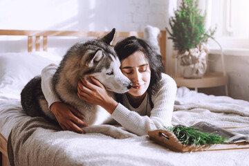 A beautiful brunette girl lies on the bed with a white husky dog at home. Sunny morning © Amina