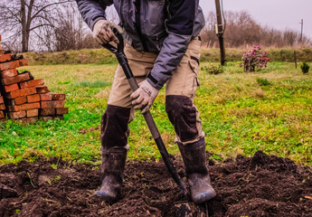 A man in a working uniform and boots with a shovel digs the ground