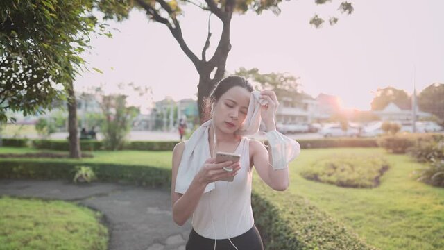 Young asian woman in sport clothing walking at the park during sunset golden hour, relaxing walk wiping sweat after hard work exercises, using phone singing to the music from earphones