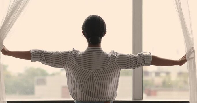 Rear view young woman standing in cozy warm apartment feels carefree and happy opening curtains let light into room, welcoming new day in the morning at home alone. Happy future, aspirations concept