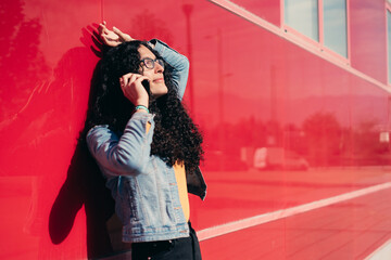 LGTB transgender girl with curly hair smiling and making a call standing on a red wall. 