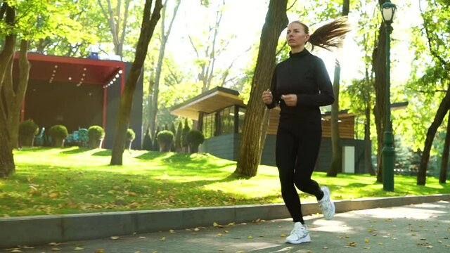 Fit woman in black sportswear jogging in park in slow motion, street cafe on background. Tracking shot of athletic female doing cardio training on sunny day. Concept of sport