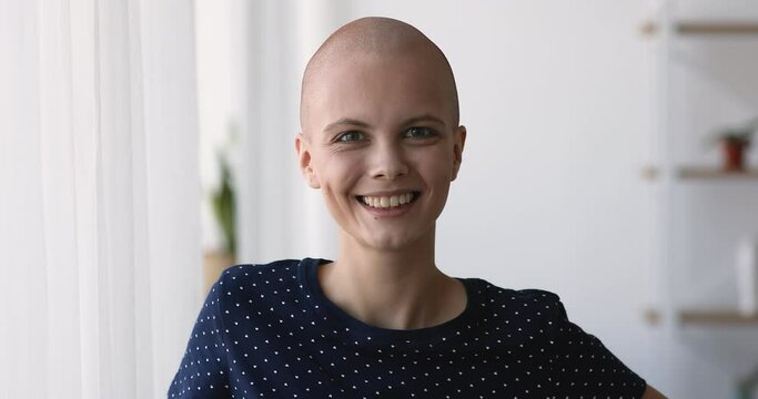 Head shot close up sincere happy young shaved head young woman looking at camera at home, feeling excited or effective cancer oncology treatment good results, hopeful about fighting disease.