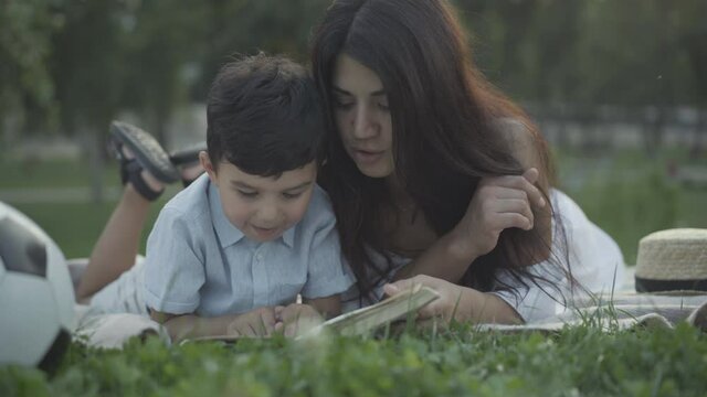 Positive Middle Eastern mother and son lying on blanket in summer park and reading book. Portrait of happy young woman and cute little boy enjoying literature at sunset. Hobby and leisure concept.