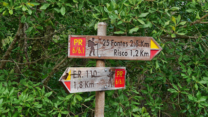 Wooden signpost with red and yellow colored markings pointing the way to the source of 25 Fontes and Risco waterfall on hiking trail on Madeira island, Portugal with dense vegetation in background.