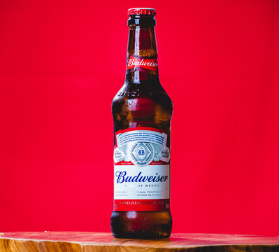 Cerveja Budweiser. Uma garrafa de cerveja da marca Budweiser sobre um objeto de madeira com plano de fundo vermelho.