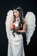 Beautiful Angel Girl offering Christmas present. Young woman with white wings in heaven style holding golden gift box on black background