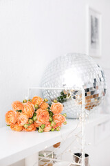 A bouquet of orange roses lies on a white shelf. Nearby is a large disco ball. Festive mood. Flowers for Christmas. Minimalism. Give flowers and gifts. New driving atmosphere. Tea peony rose.