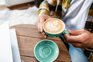 cropped of man sitting in cafe and holding cup of coffee with latte art