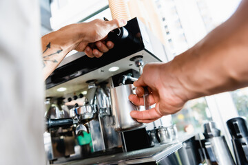 partial of barista holding metallic milk mug near coffee machine steamer on blurred foreground