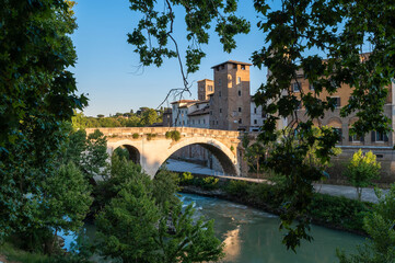 Fototapeta na wymiar Rome Isola Tiberina, Ponte Fabricio from the Lungotevere dei Cenci, called the bridge of the 4 heads in travertine formed by large arches. Beautiful reflections of the luxuriant tiber vegetation.