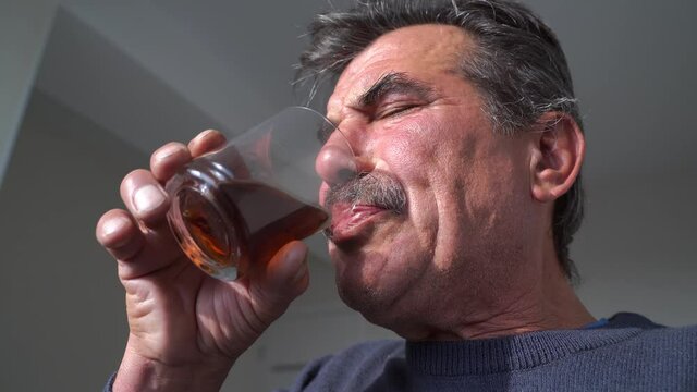From below middle aged man with shaking hand sipping alcohol and wincing at hom