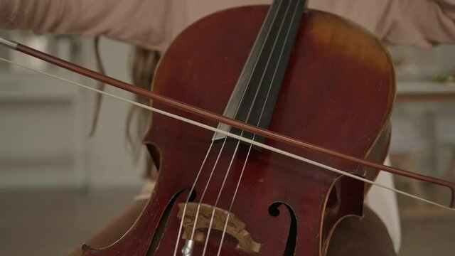 Closeup of young woman hands playing cello sitting on the chair at home interior. Light minimalist room with musician woman training cello holding a bow, gimbal shot