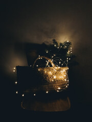 Rustic basket with Christmas tree branches in lights on wooden chair in the dark. Aesthetic holiday