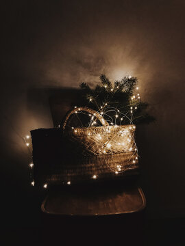 Rustic basket with Christmas tree branches in lights on wooden chair in the dark. Aesthetic holiday