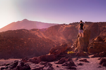 A young hiker wearing a black t-shirt and purple shorts looking at the Teide volcano mounted on a stone surronded by a red, volcanic landscape. Teide National Park, Tenerife, Canary Islands, Spain.