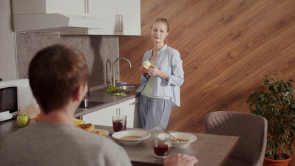 In the morning, in the kitchen, the guy has breakfast at the table, the girl removes the peel from the apple and eats it